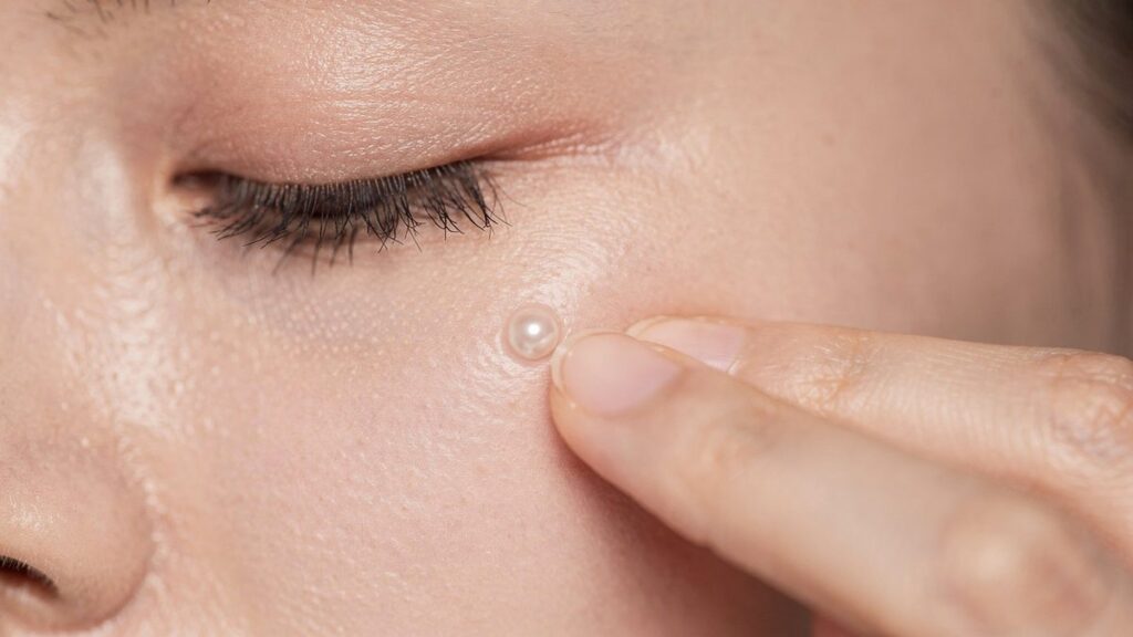 Dedo anular aplicando una perla de crema de contorno bajo el ojo cerrado